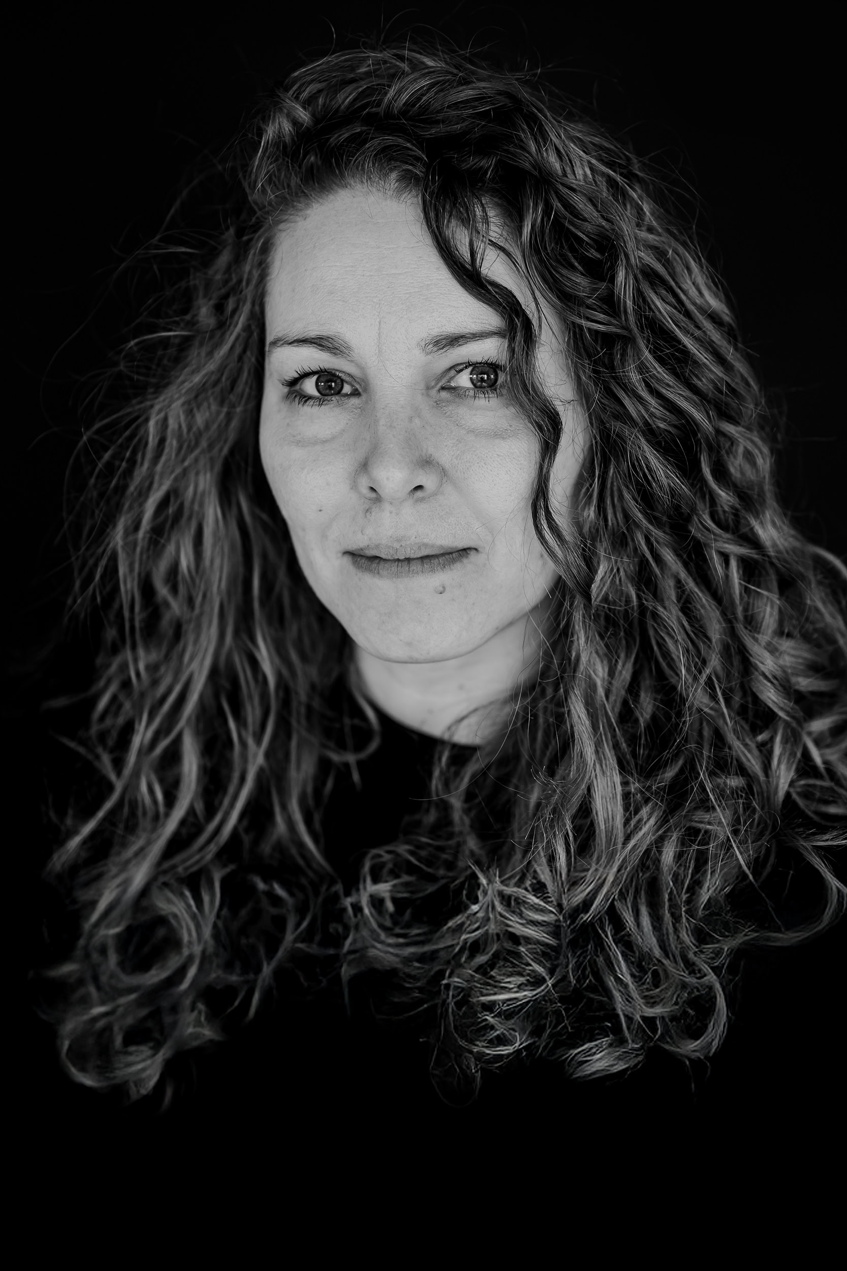 Das Bild zeigt eine Frau mit lockigem, vollem Haar, die direkt in die Kamera blickt, was eine kraftvolle und authentische Ausstrahlung vermittelt. Fotografiert von Mica Zeitz, strahlt das Portrait eine intime Boudoir-Atmosphäre aus, die die natürliche Schönheit der Person betont.